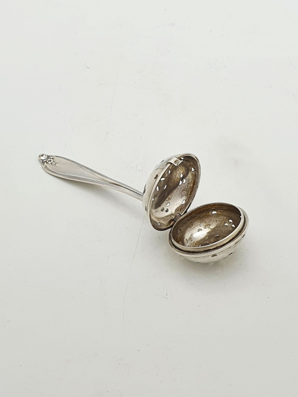 A George V sterling silver hinged tea infuser spoon featuring a perforated spherical bowl attached to a decorated handle. View II