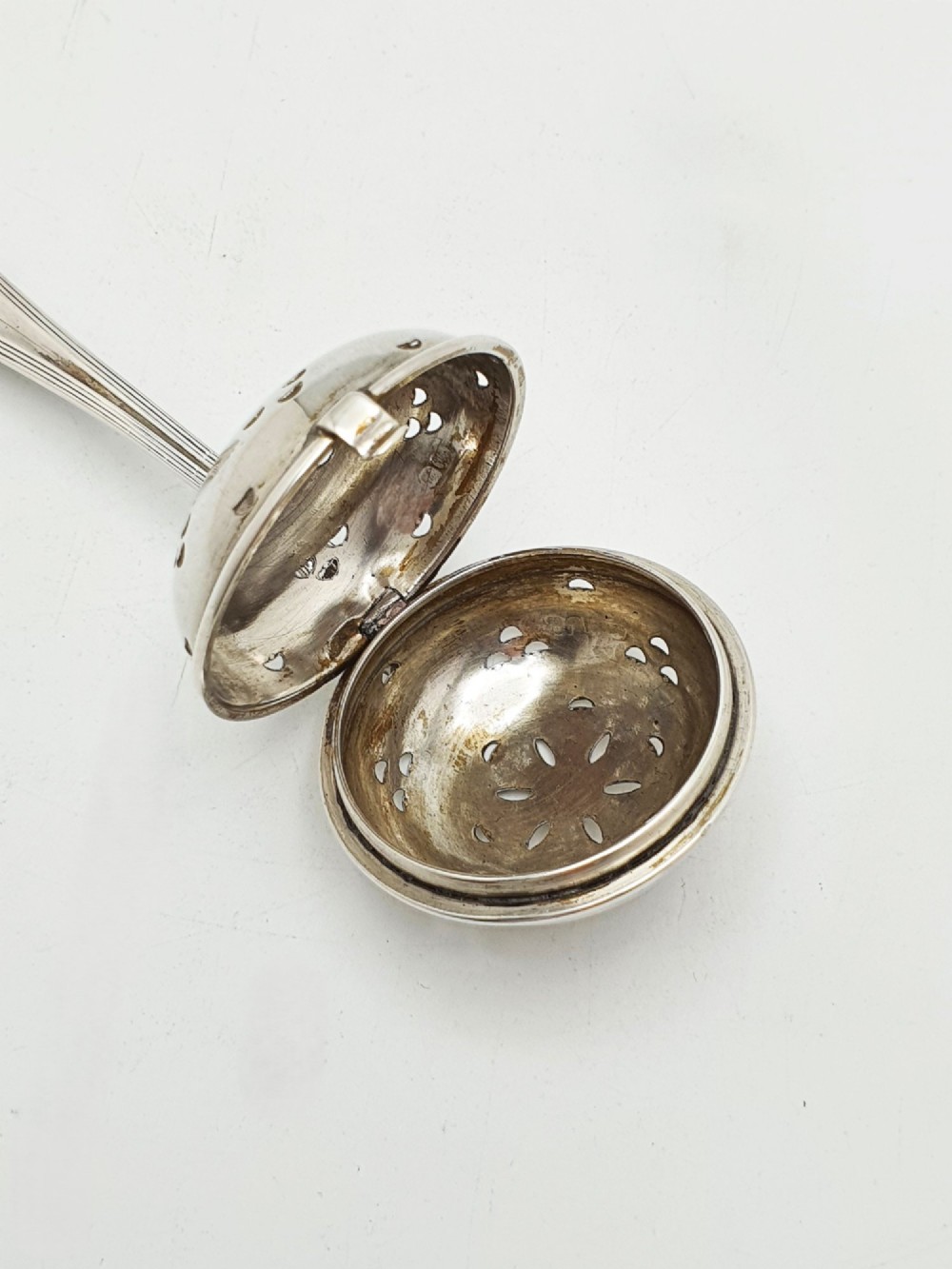 A George V sterling silver hinged tea infuser spoon featuring a perforated spherical bowl attached to a decorated handle. View IV