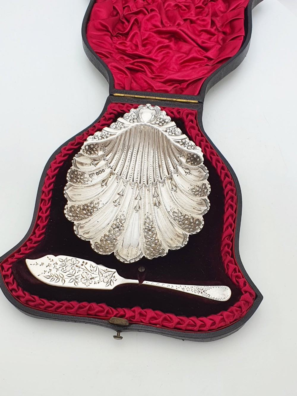 A Victorian sterling silver scallop shell butter dish and matching engraved spreader knife resting inside a fitted red and black velvet presentation case.