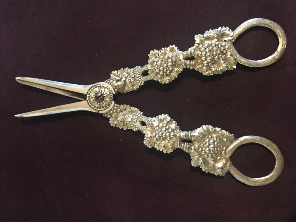 A pair of silver grape scissors featuring handles relief-moulded with grape clusters and vine leaves leading to round finger loops.