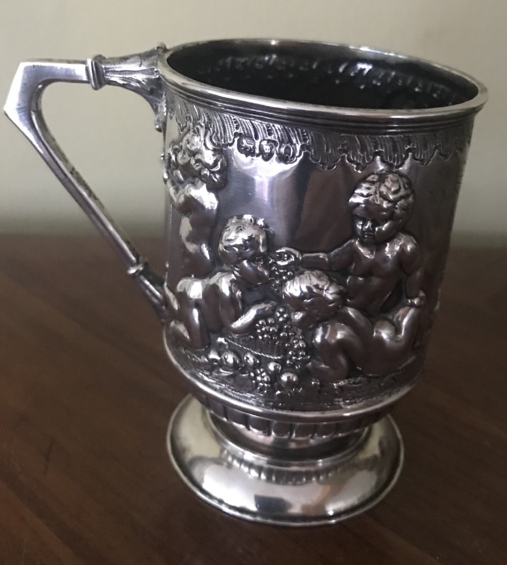 A silver mug decorated with repousse cherubs in relief, featuring an angular scroll handle and a stepped pedestal foot. View V