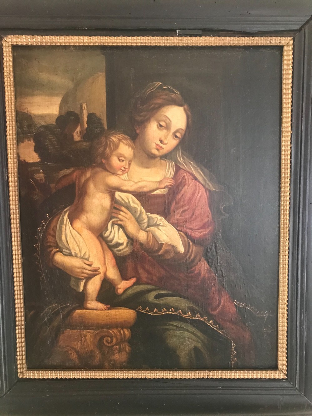 An antique oil painting depicting the Madonna in a burgundy dress holding the infant Christ, set within a heavy ebonised wooden moulding frame. View II