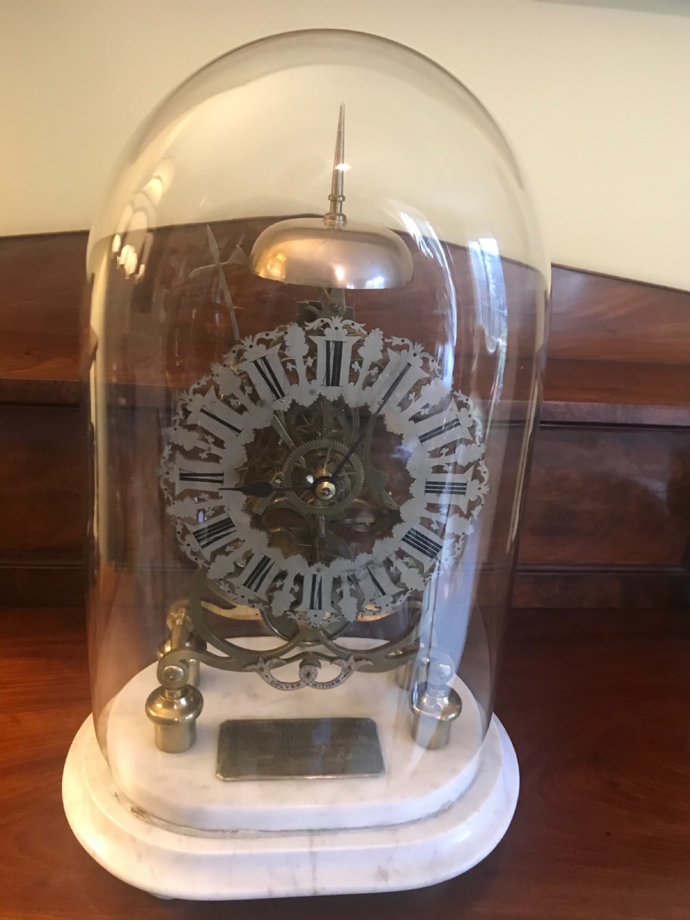 A Victorian brass skeleton clock with an exposed gear mechanism and silvered Roman numeral dial, encased in a clear glass dome on a white marble base.