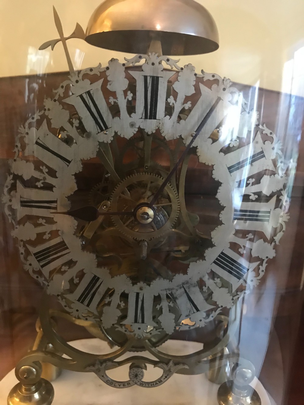 A Victorian brass skeleton clock with an exposed gear mechanism and silvered Roman numeral dial, encased in a clear glass dome on a white marble base. View II