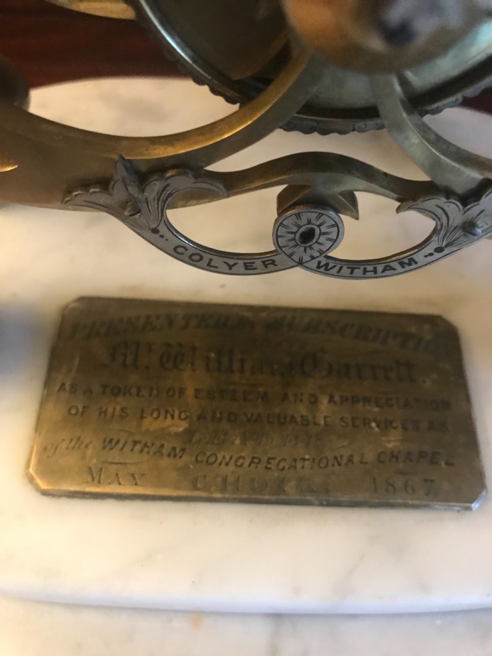 A Victorian brass skeleton clock with an exposed gear mechanism and silvered Roman numeral dial, encased in a clear glass dome on a white marble base. View IV