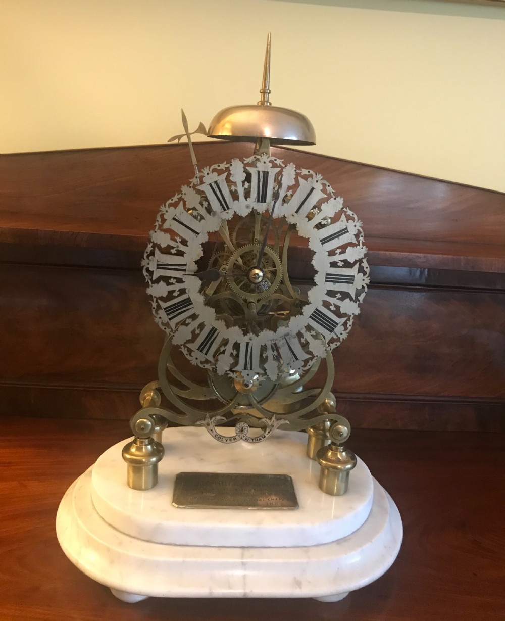 A Victorian brass skeleton clock with an exposed gear mechanism and silvered Roman numeral dial, encased in a clear glass dome on a white marble base. View V