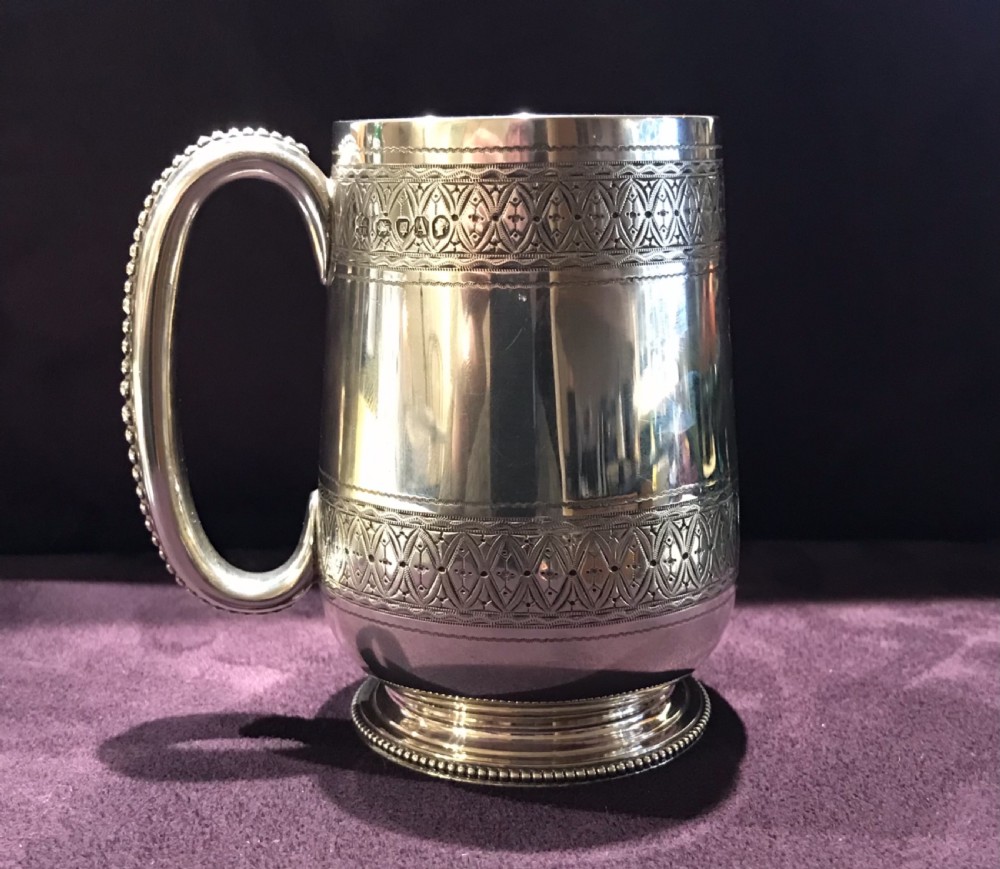 An antique sterling silver mug featuring engraved geometric borders, a beaded handle, and hallmarked stamps near the top rim.