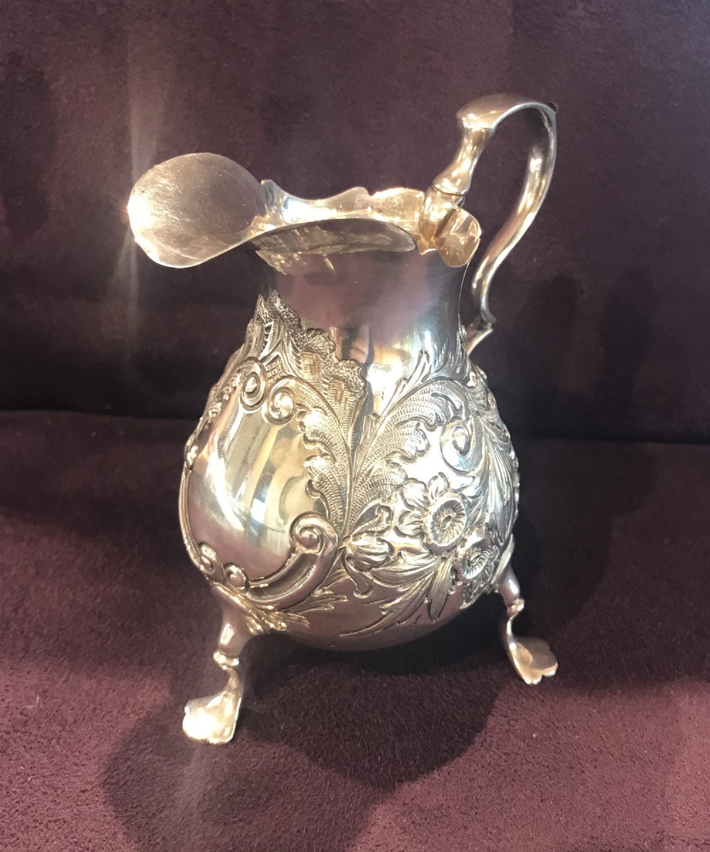 An antique silver cream jug featuring embossed floral motifs, a double-scroll handle, and three pad feet.