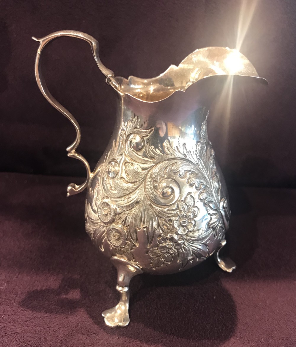An antique silver cream jug featuring embossed floral motifs, a double-scroll handle, and three pad feet. View II