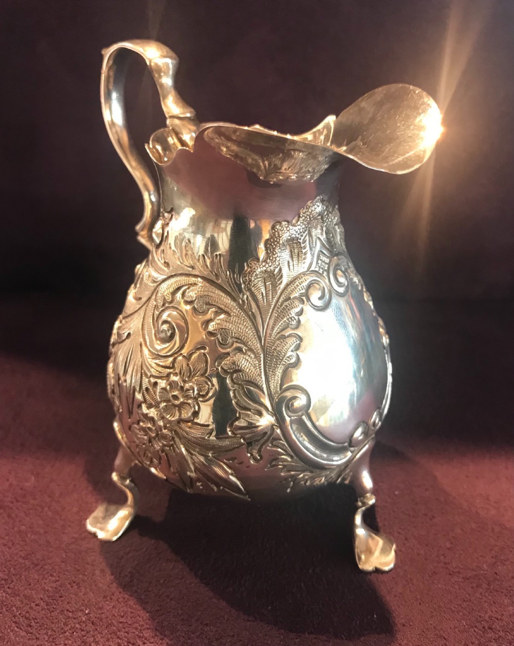 An antique silver cream jug featuring embossed floral motifs, a double-scroll handle, and three pad feet. View V
