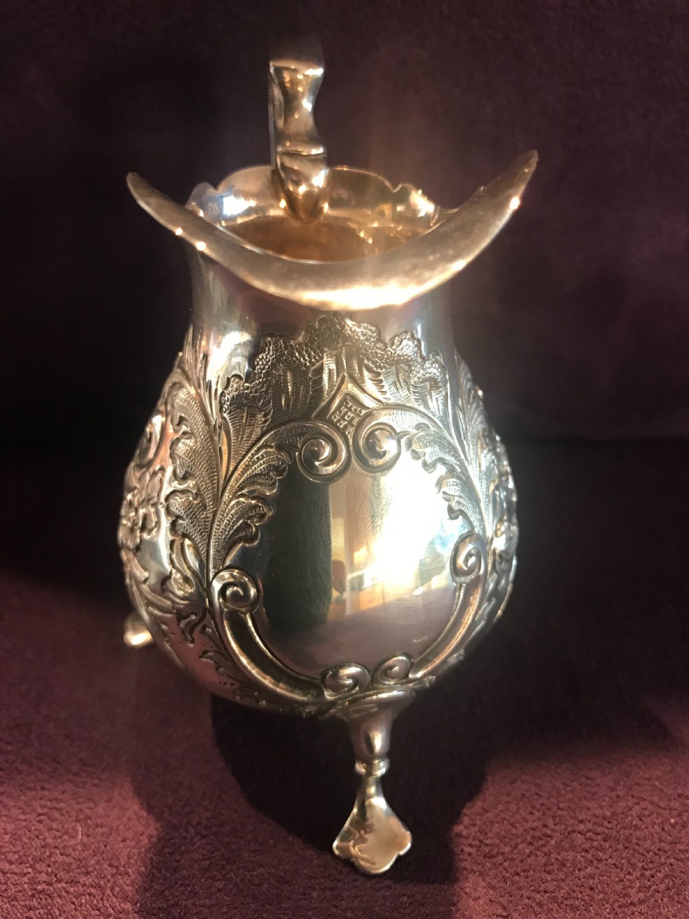 An antique silver cream jug featuring embossed floral motifs, a double-scroll handle, and three pad feet. View VI