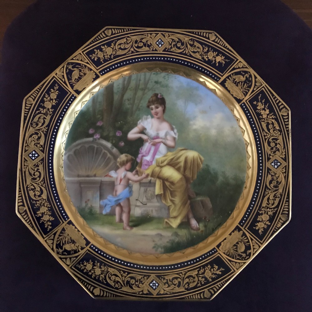 An octagonal antique porcelain cabinet plate with a cobalt blue and gold gilded border surrounding a central hand-painted scene of a classical woman and cherub.