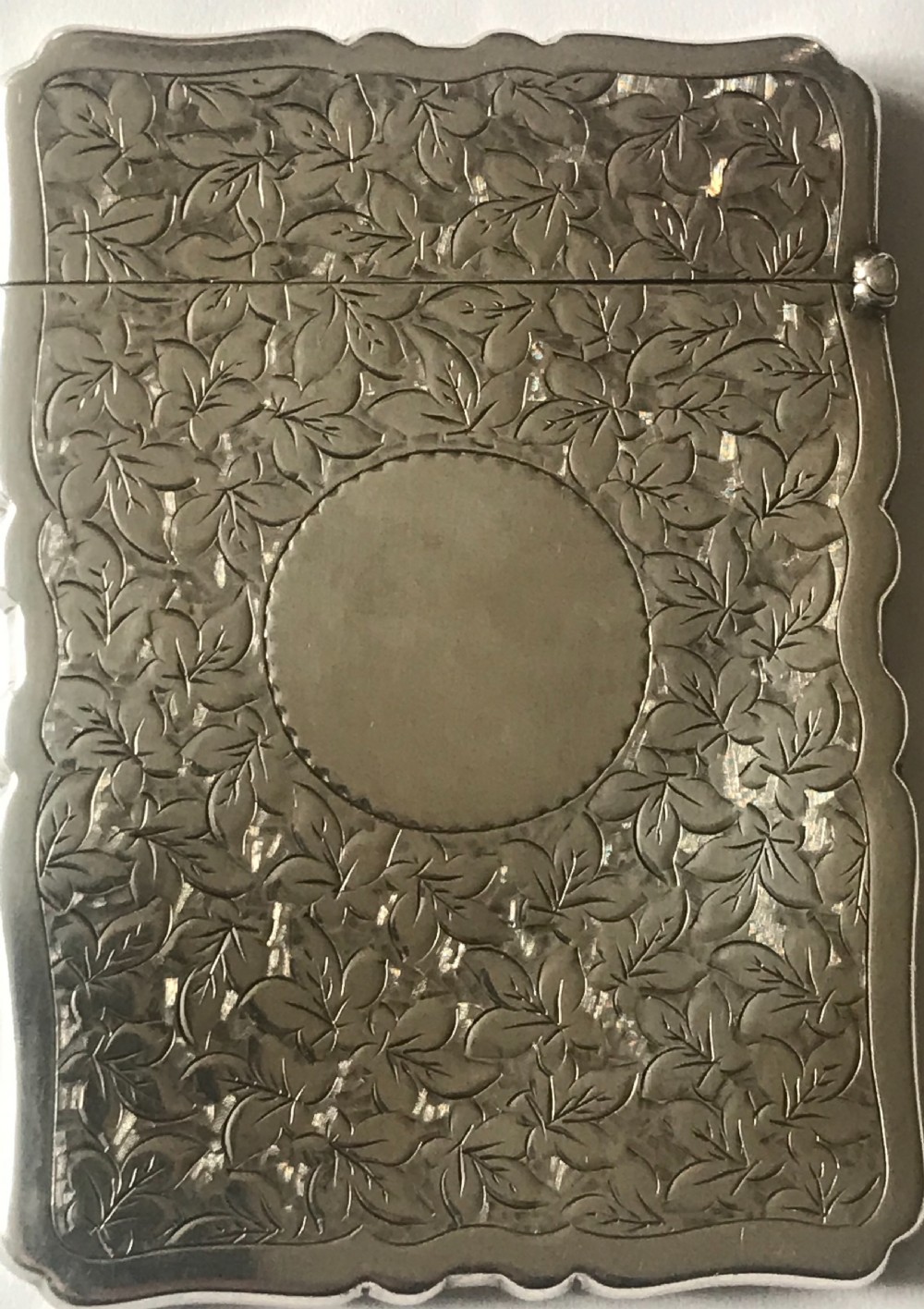 An antique silver card case featuring engraved leaf motifs, shaped scalloped edges, and a central circular cartouche.