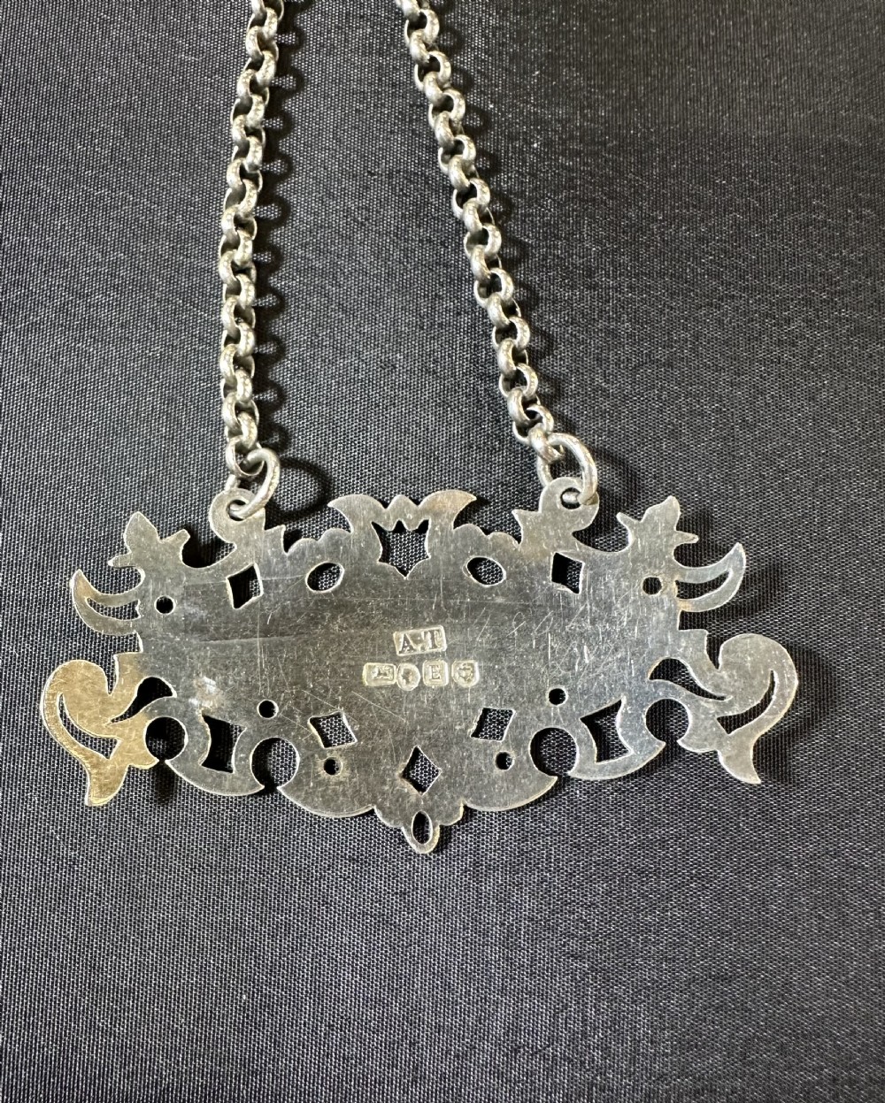 A Victorian sterling silver decanter label featuring pierced scrollwork and stamped British hallmarks suspended from a silver chain.