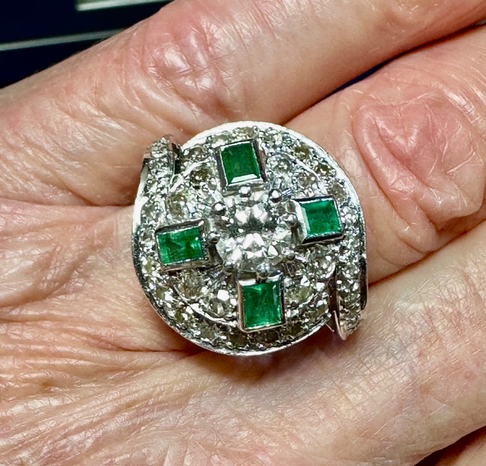 An Art Deco style 18 carat white gold cocktail ring featuring a central diamond surrounded by rectangular green emeralds and pave-set diamonds in a swirling geometric design. View II