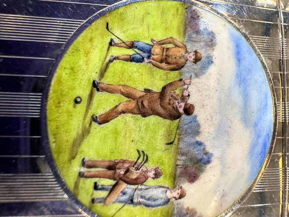 An engine-turned sterling silver vesta case featuring a central round enamelled plaque depicting three golfers on a golf course. View V