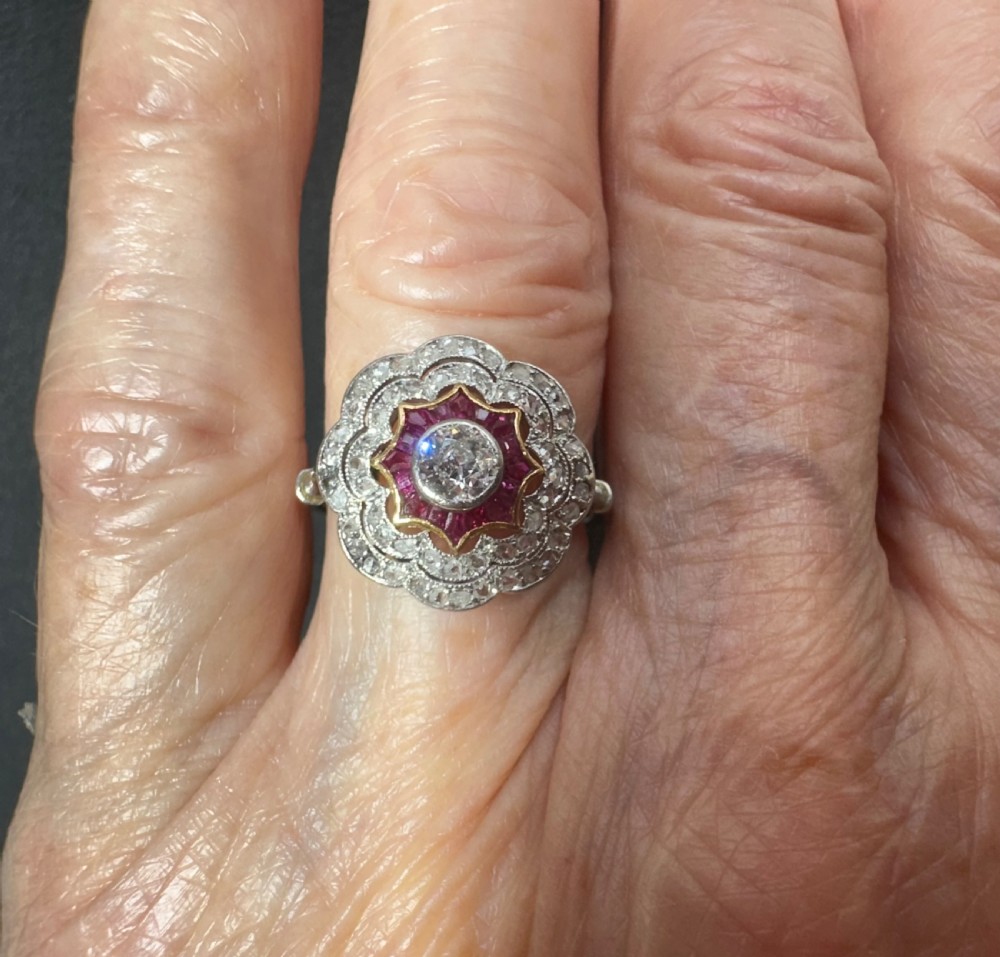 An Edwardian platinum cluster ring featuring a central round diamond encircled by a star-shaped border of calibre-cut red rubies and a scalloped diamond halo. View II