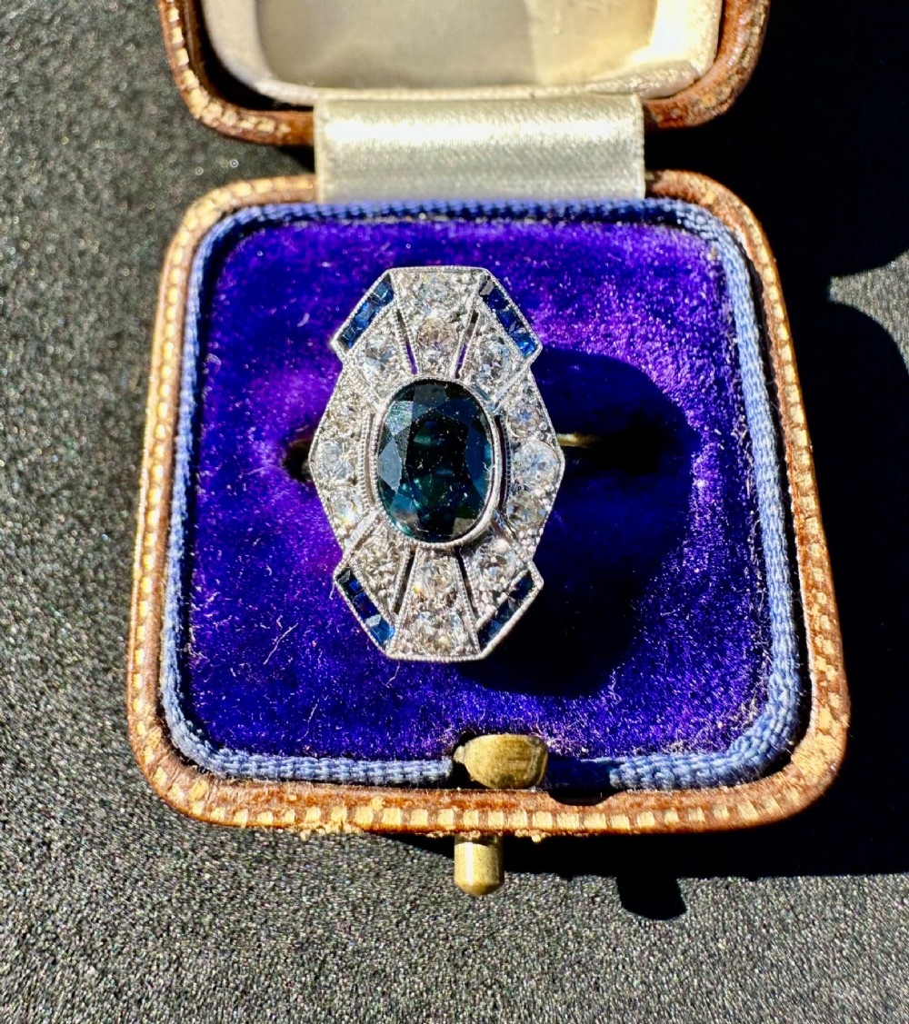 An Art Deco geometric panel ring featuring a central oval blue sapphire surrounded by diamonds and calibre-cut sapphire accents in a white metal and gold setting.