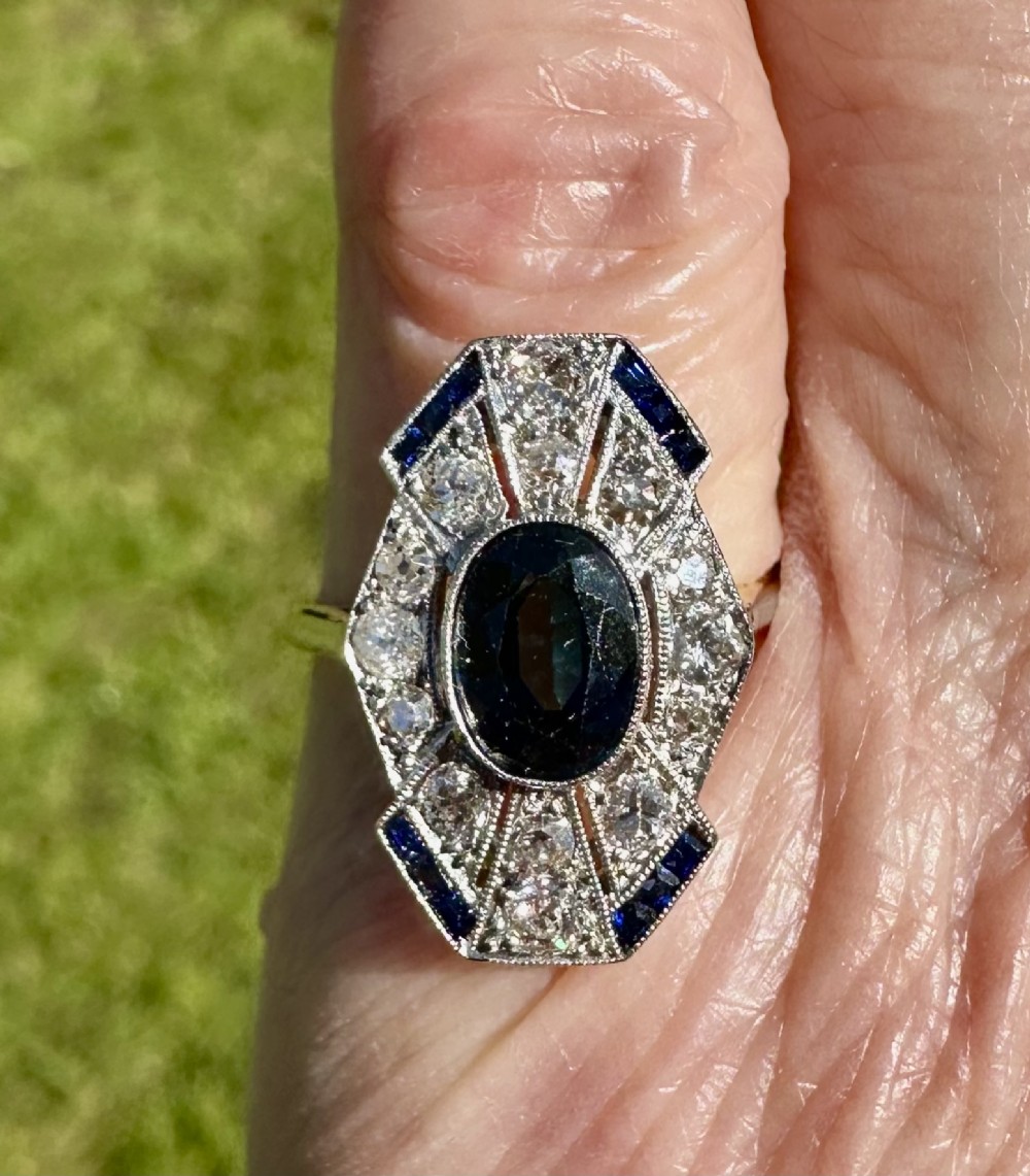 An Art Deco geometric panel ring featuring a central oval blue sapphire surrounded by diamonds and calibre-cut sapphire accents in a white metal and gold setting. View II