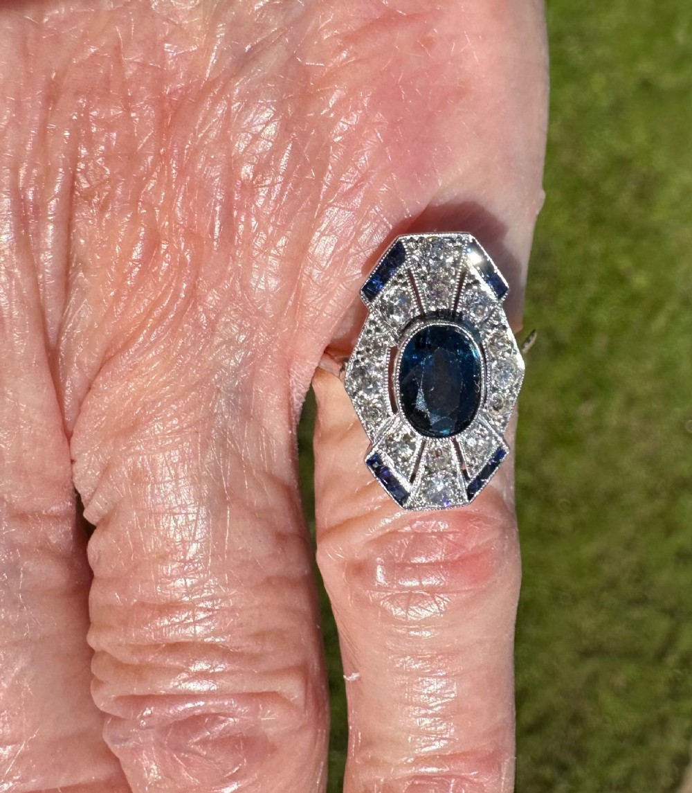 An Art Deco geometric panel ring featuring a central oval blue sapphire surrounded by diamonds and calibre-cut sapphire accents in a white metal and gold setting. View III