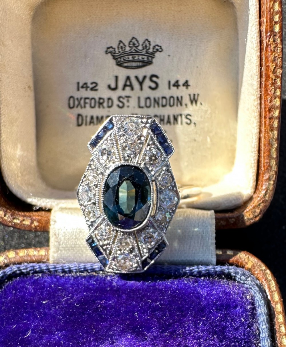 An Art Deco geometric panel ring featuring a central oval blue sapphire surrounded by diamonds and calibre-cut sapphire accents in a white metal and gold setting. View V