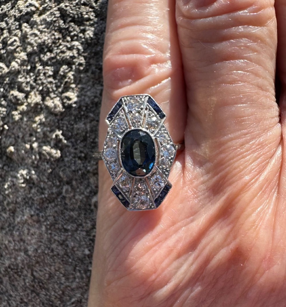 An Art Deco geometric panel ring featuring a central oval blue sapphire surrounded by diamonds and calibre-cut sapphire accents in a white metal and gold setting. View VI
