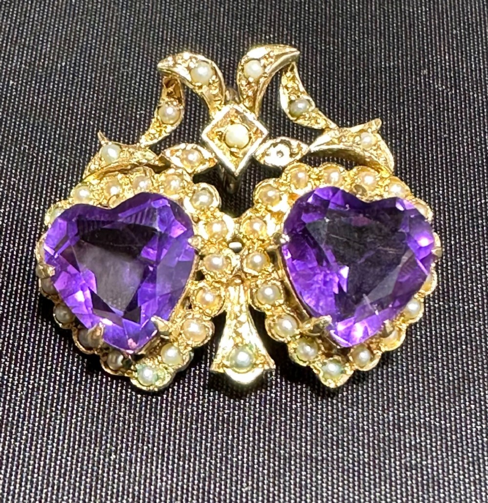 An antique 9ct gold double heart brooch set with two faceted purple amethysts surrounded by seed pearls and topped with a ribbon bow motif.