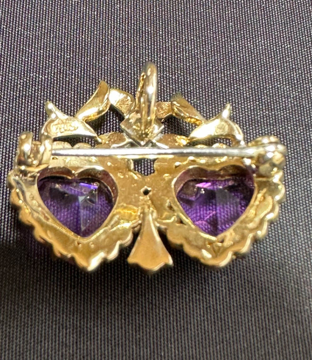 An antique 9ct gold double heart brooch set with two faceted purple amethysts surrounded by seed pearls and topped with a ribbon bow motif. View II