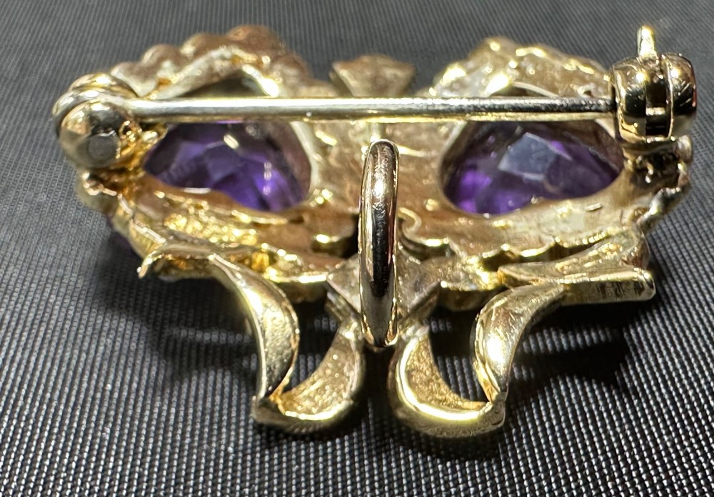 An antique 9ct gold double heart brooch set with two faceted purple amethysts surrounded by seed pearls and topped with a ribbon bow motif. View III