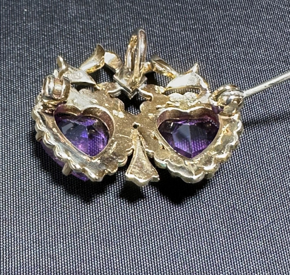 An antique 9ct gold double heart brooch set with two faceted purple amethysts surrounded by seed pearls and topped with a ribbon bow motif. View IV