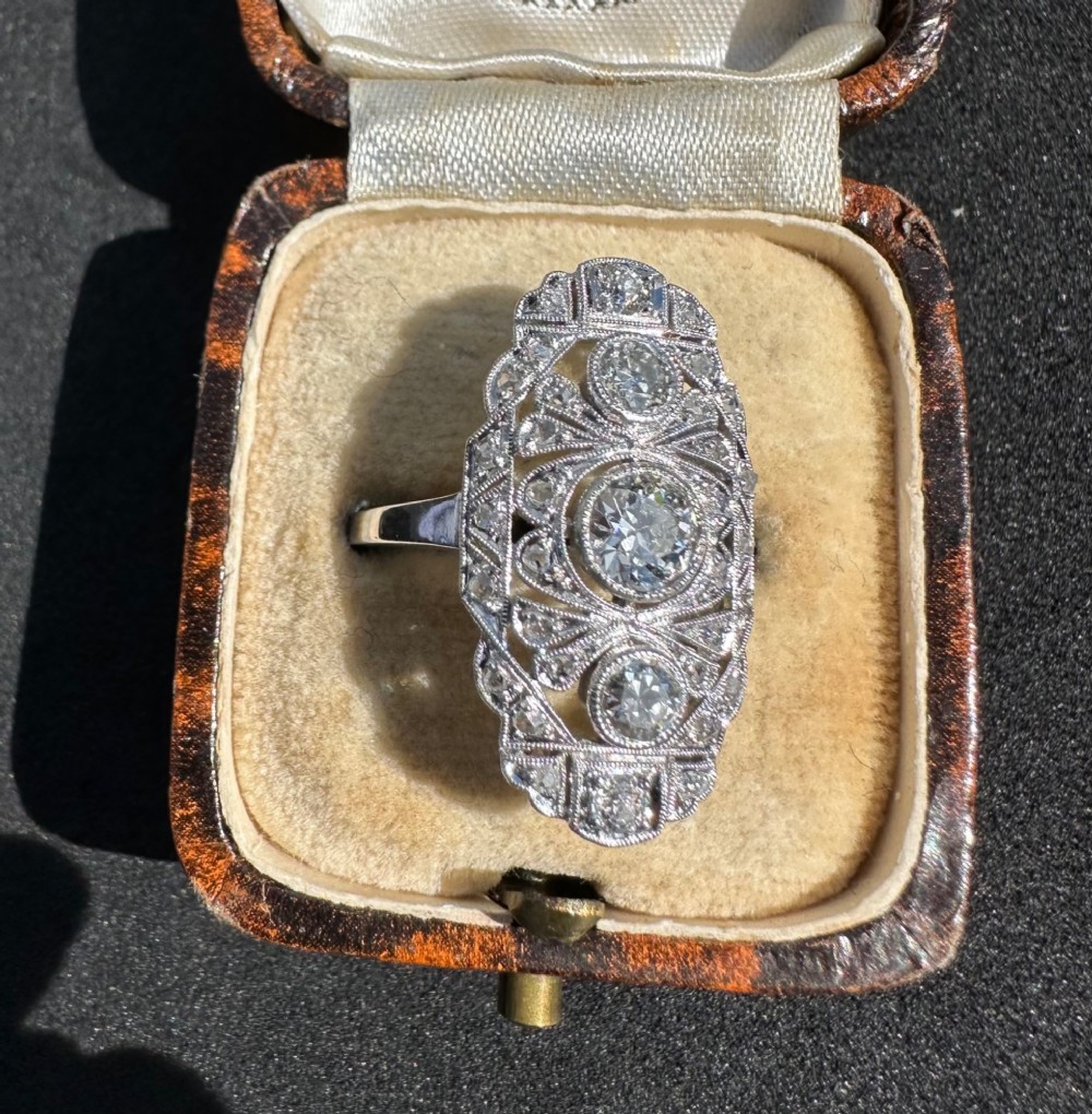 An Art Deco platinum panel ring featuring an elongated geometric openwork design set with multiple circular diamonds and milgrain detailing.