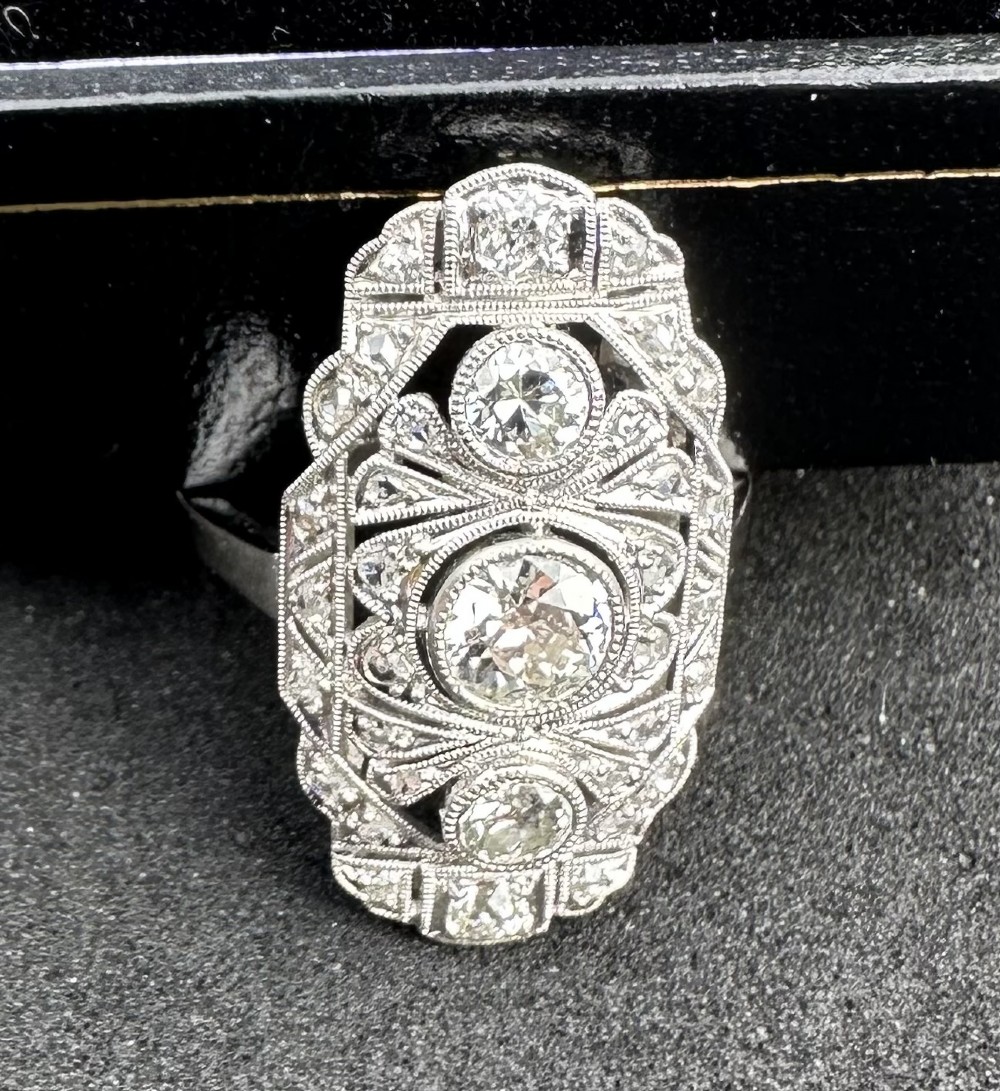 An Art Deco platinum panel ring featuring an elongated geometric openwork design set with multiple circular diamonds and milgrain detailing. View V