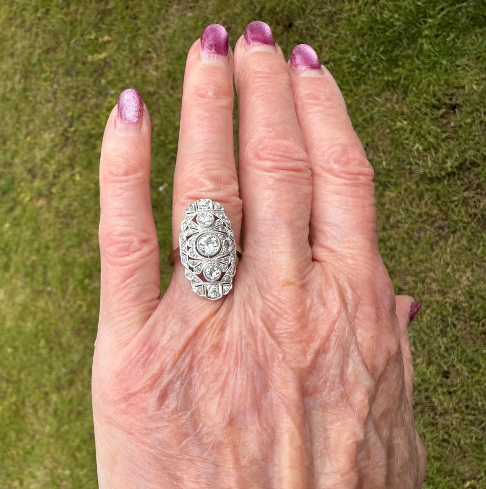 An Art Deco platinum panel ring featuring an elongated geometric openwork design set with multiple circular diamonds and milgrain detailing. View VII