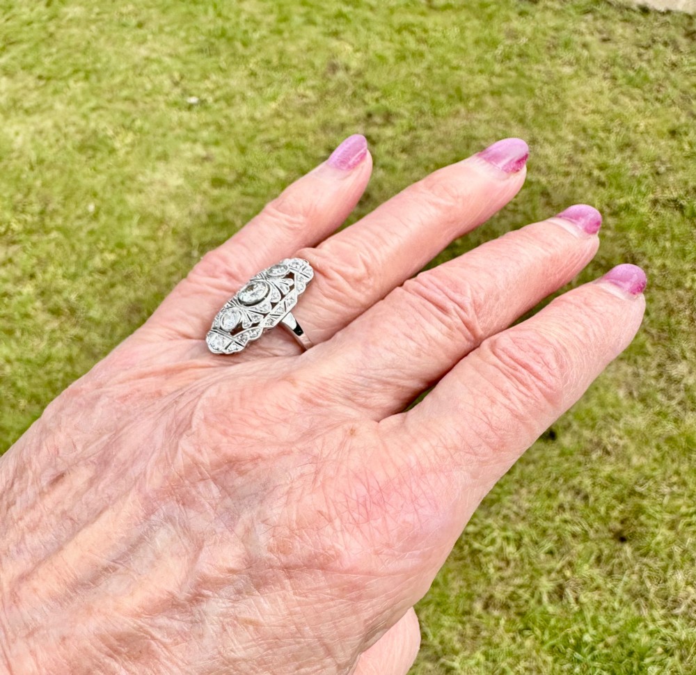 An Art Deco platinum panel ring featuring an elongated geometric openwork design set with multiple circular diamonds and milgrain detailing. View VIII