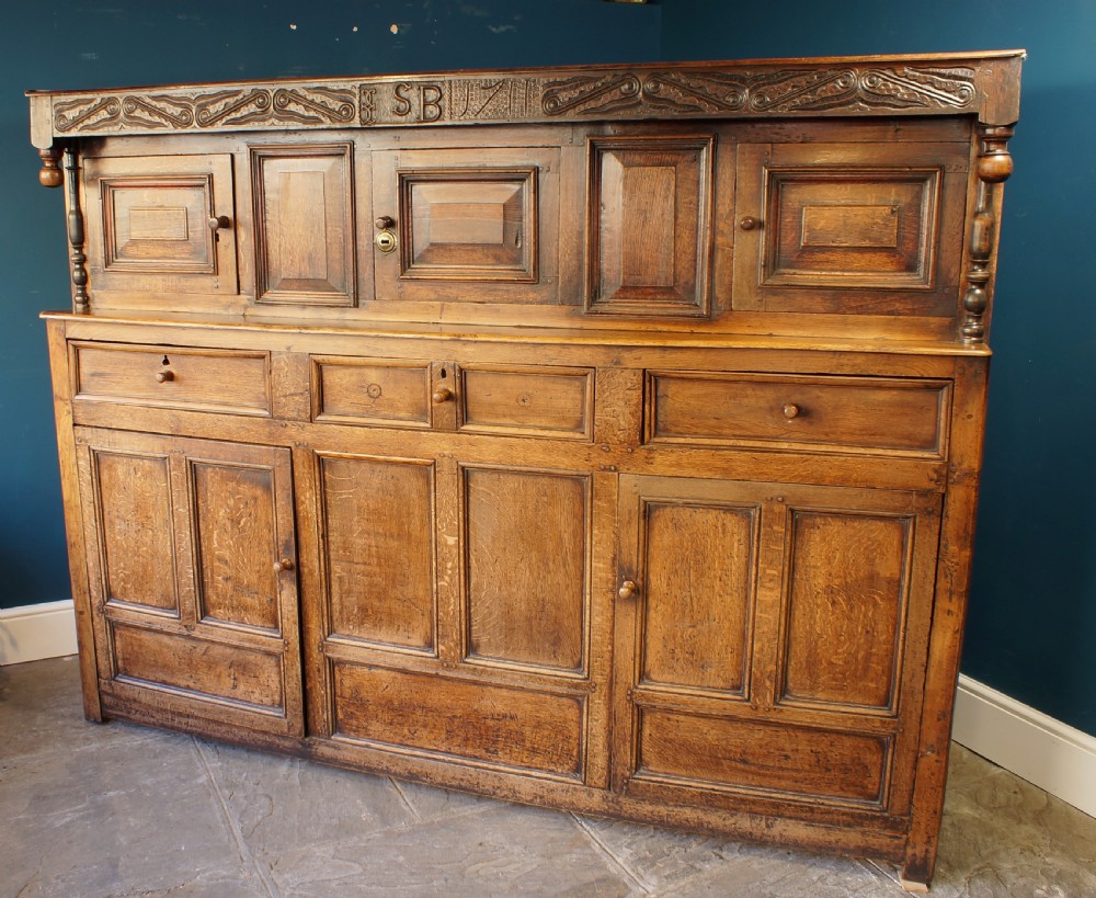 A large 18th-century carved oak court cupboard with recessed upper doors, carved top frieze with initials, and three lower cupboard doors.