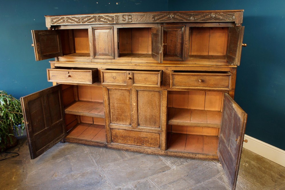 A large 18th-century carved oak court cupboard with recessed upper doors, carved top frieze with initials, and three lower cupboard doors. View XI