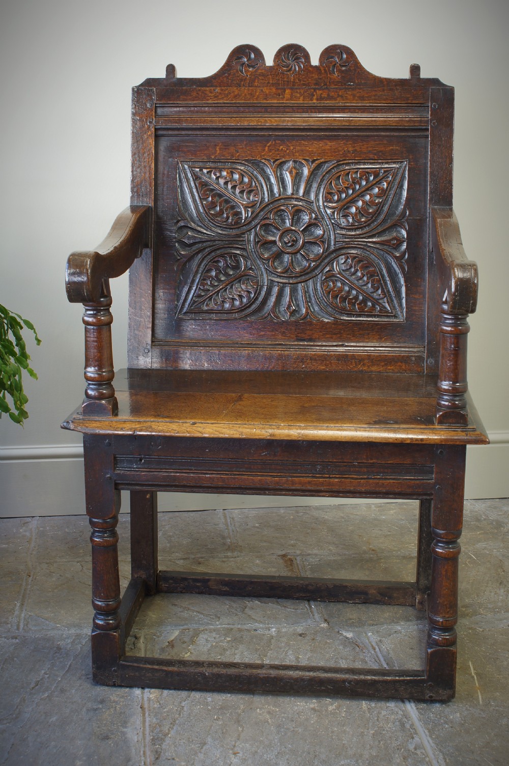A 17th-century dark oak wainscot armchair with a deeply carved back panel, turned front legs, and sturdy box stretchers.