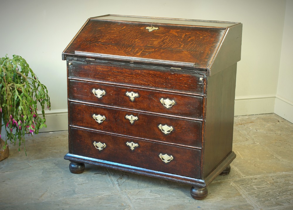 A William and Mary dark oak slant-front bureau featuring geometric parquetry on the lid, three long drawers with brass drop handles, and bun feet.