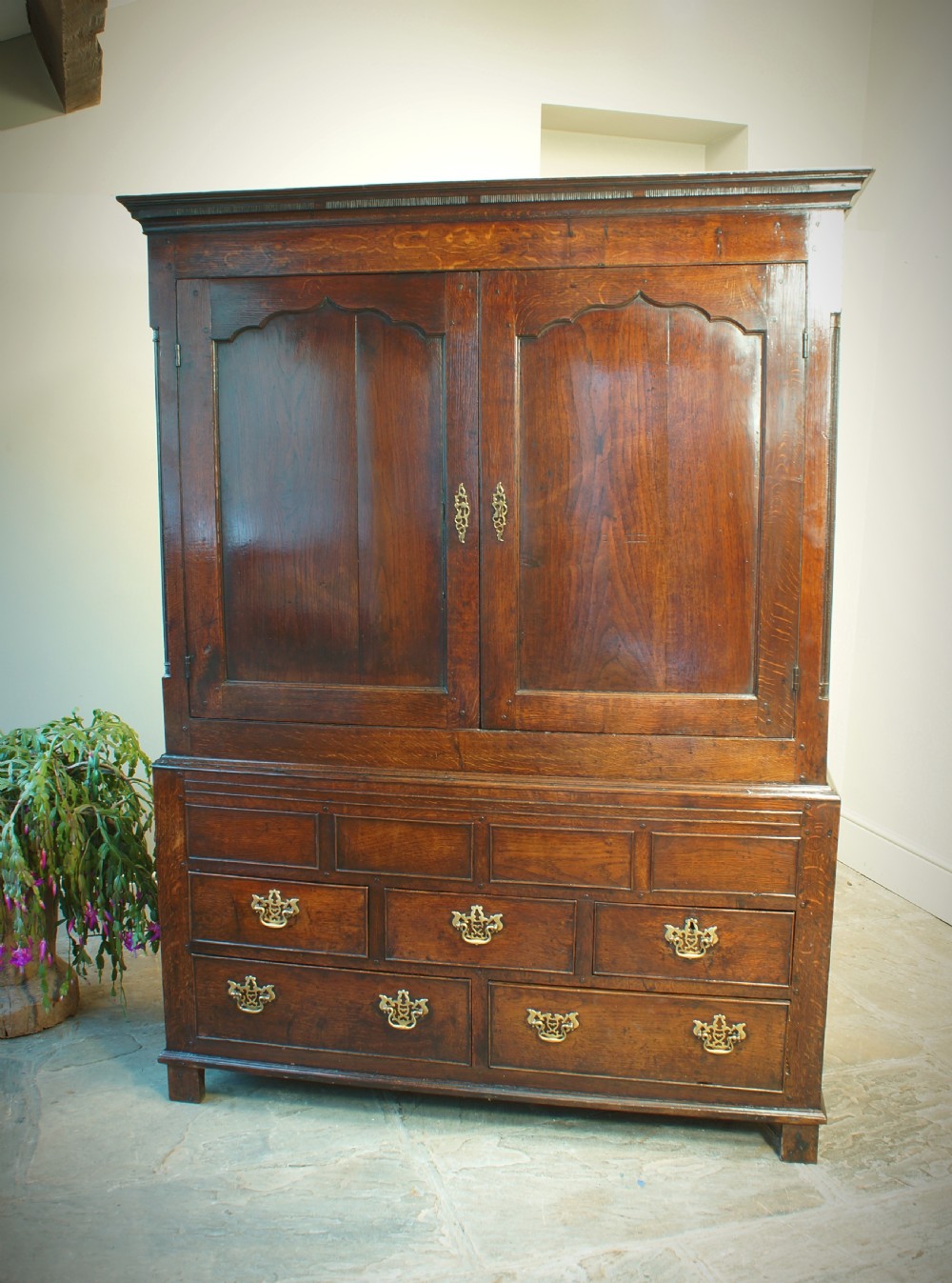 A Georgian oak linen press featuring two arched upper panel doors over a lower chest section with decorative brass hardware.