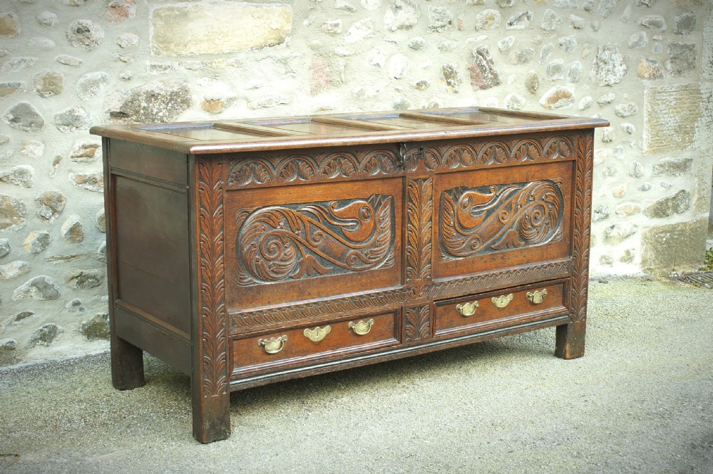 A 17th century dark oak mule chest featuring carved scrollwork front panels, two lower drawers with brass handles, and a panelled top lid.