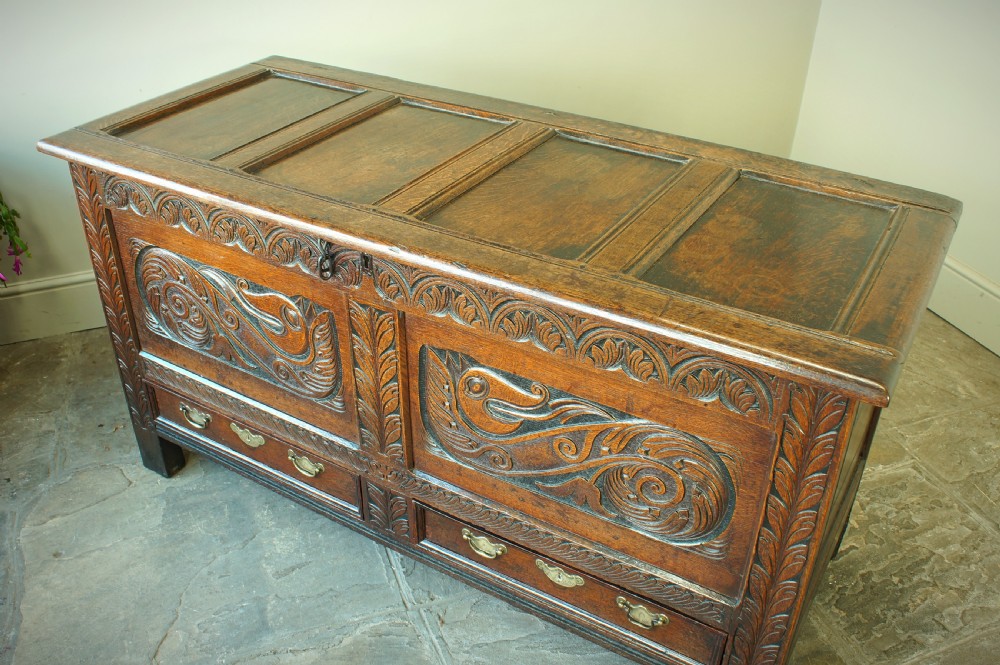 A 17th century dark oak mule chest featuring carved scrollwork front panels, two lower drawers with brass handles, and a panelled top lid. View V