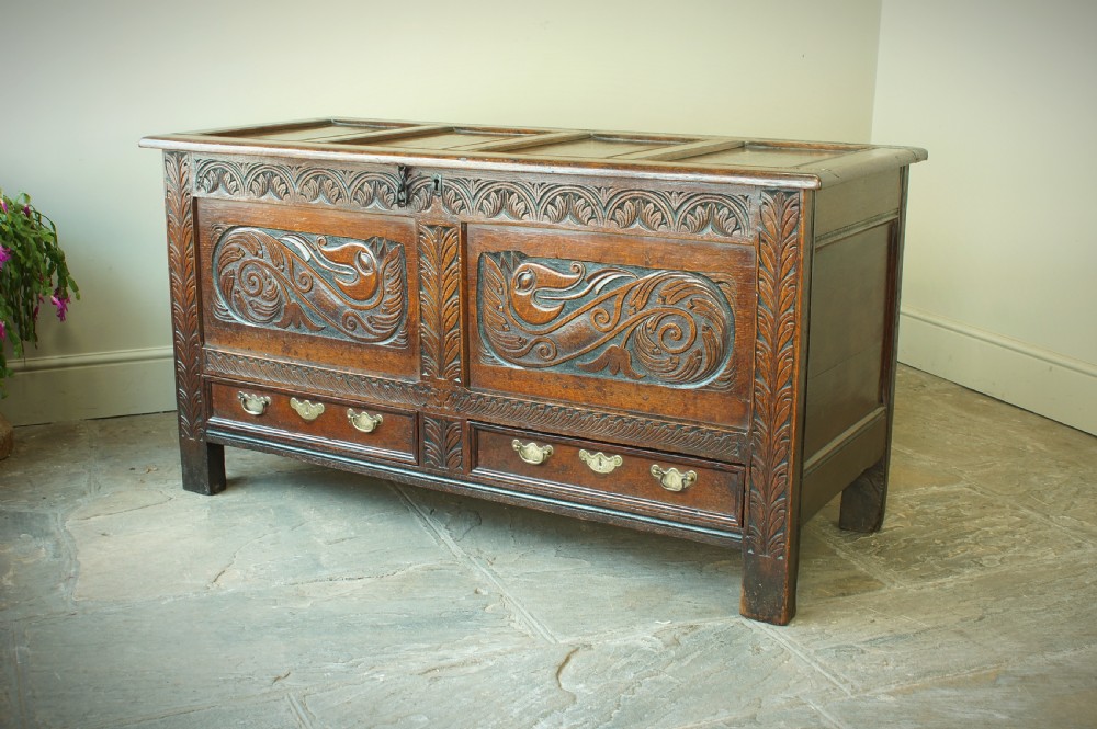 A 17th century dark oak mule chest featuring carved scrollwork front panels, two lower drawers with brass handles, and a panelled top lid. View VII