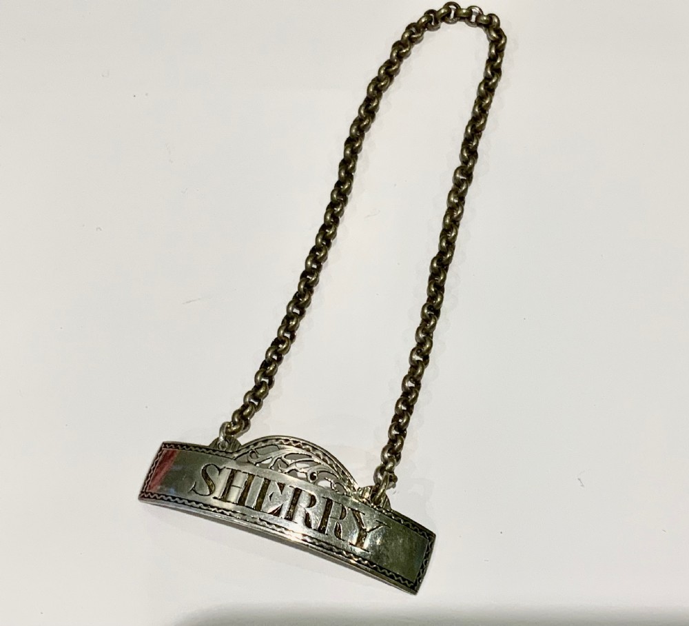 An antique Georgian sterling silver decanter label inscribed with SHERRY, featuring an engraved border, pierced crest, and attached chain.
