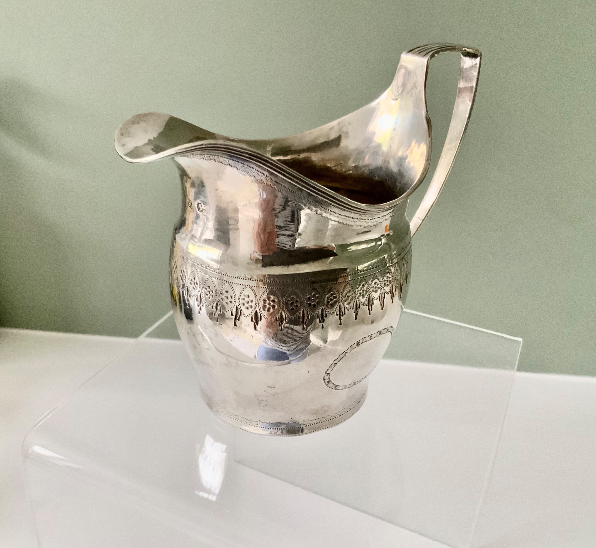 A Georgian sterling silver cream jug with bright-cut engraving, a broad spout, an unengraved oval cartouche, and a shaped handle.