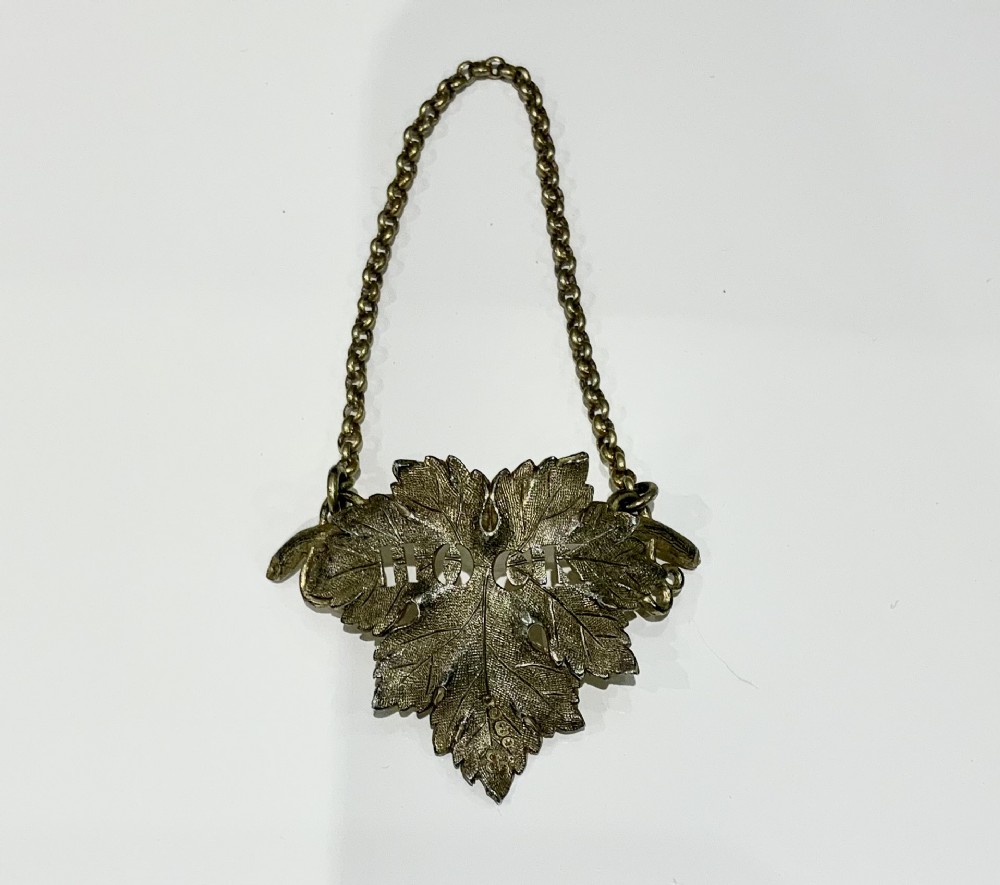 A silver gilt decanter label shaped as a detailed vine leaf with pierced lettering reading HOCK, suspended by a chain.