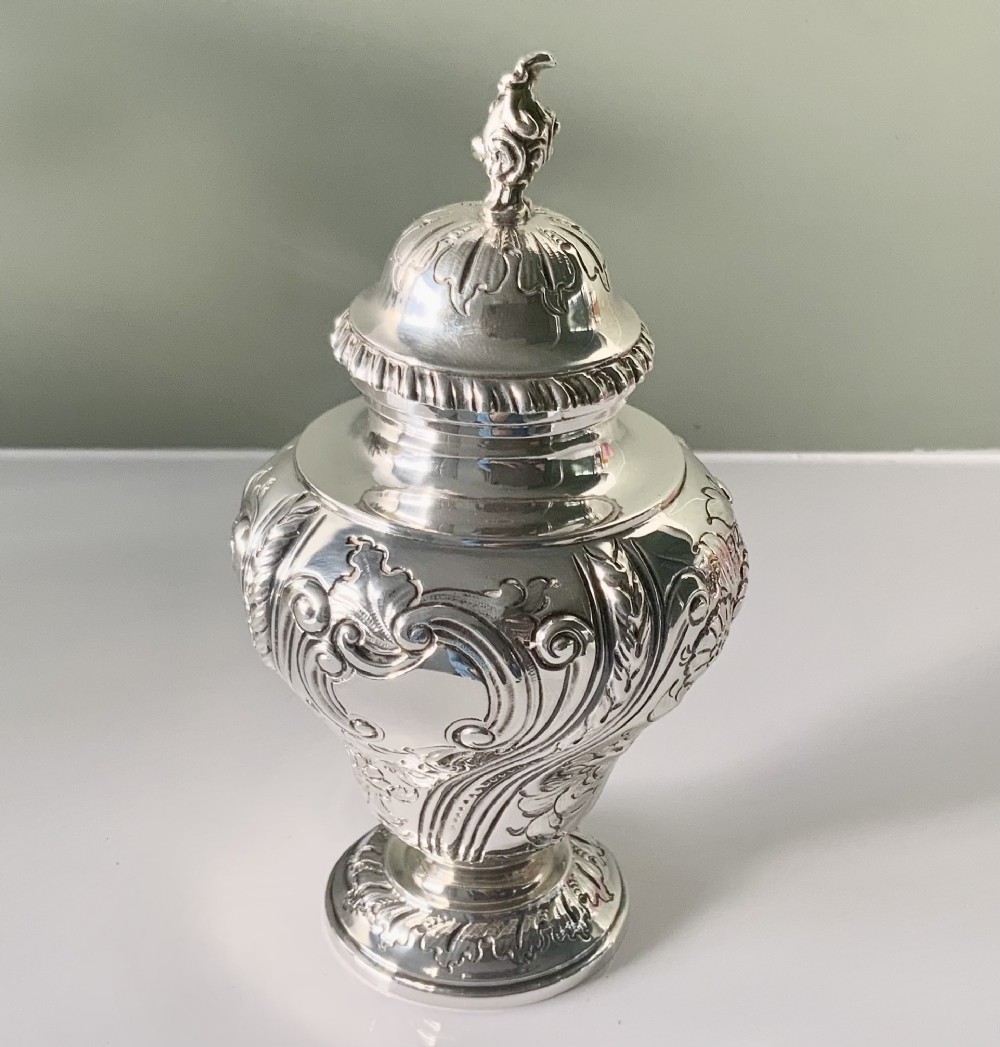 An ornate silver tea caddy featuring embossed scrollwork, a pedestal base, and a decorative finial on the lid.