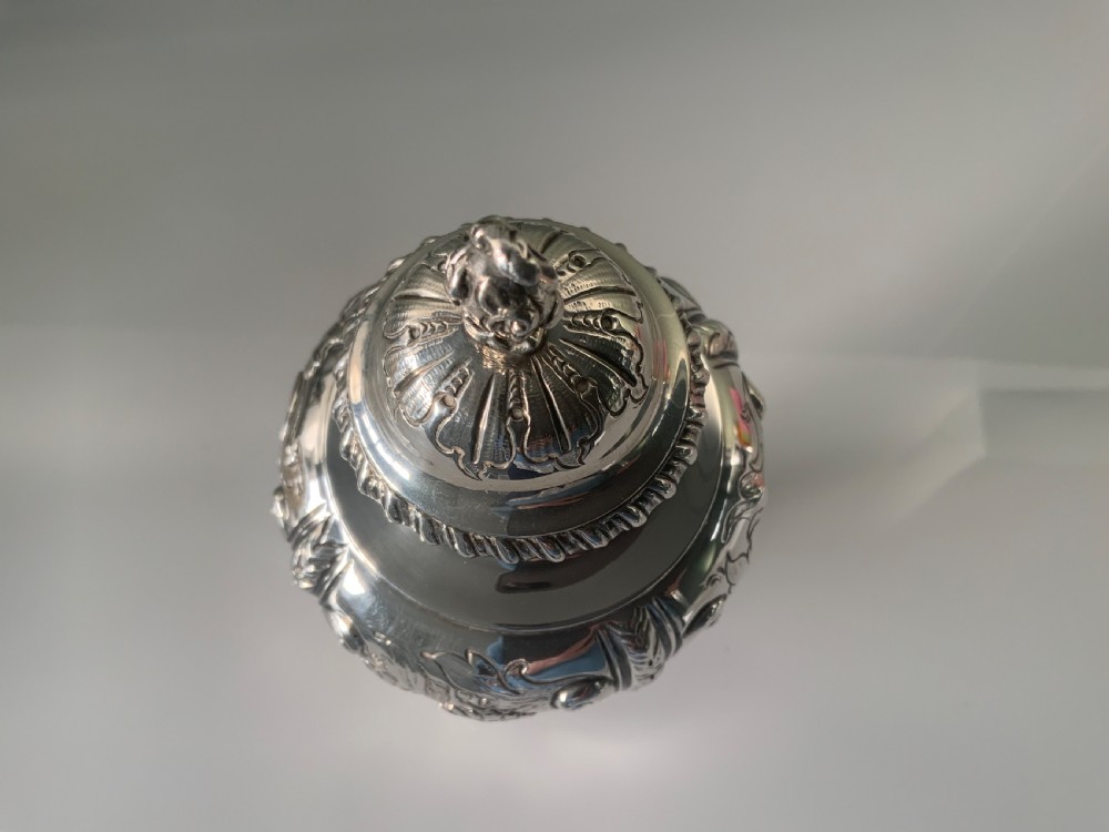 An ornate silver tea caddy featuring embossed scrollwork, a pedestal base, and a decorative finial on the lid. View V