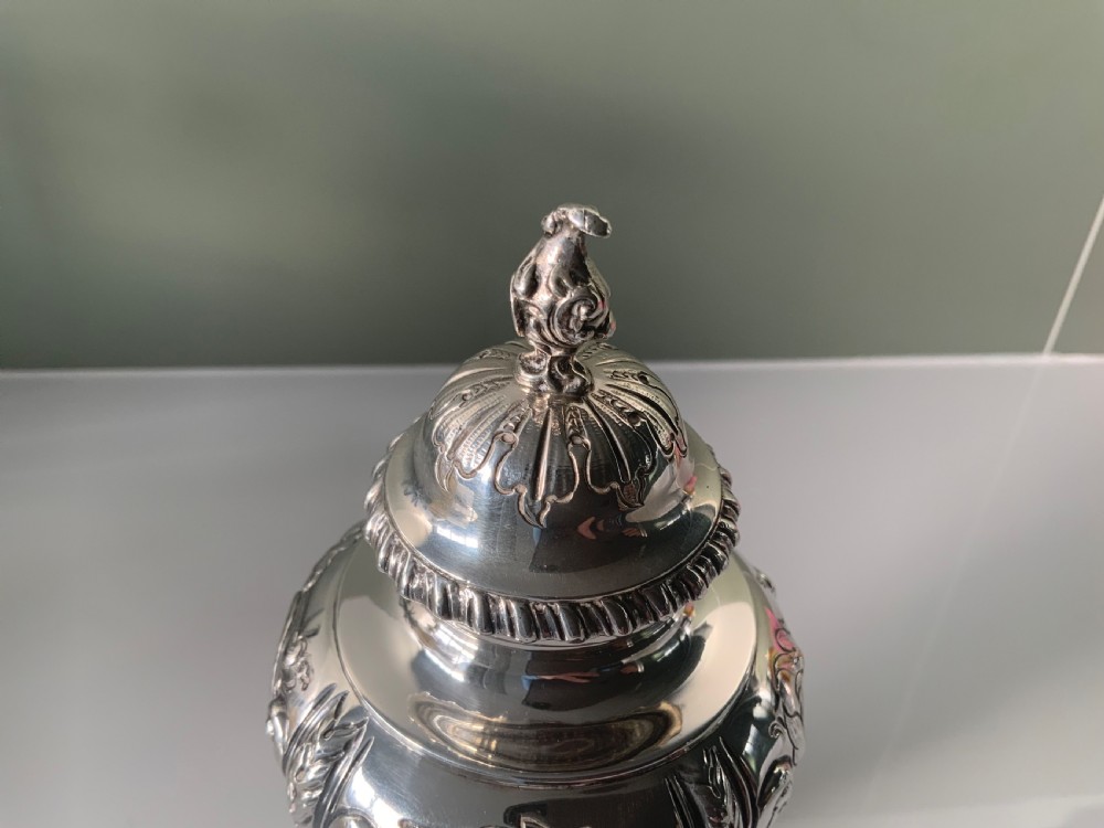 An ornate silver tea caddy featuring embossed scrollwork, a pedestal base, and a decorative finial on the lid. View VI
