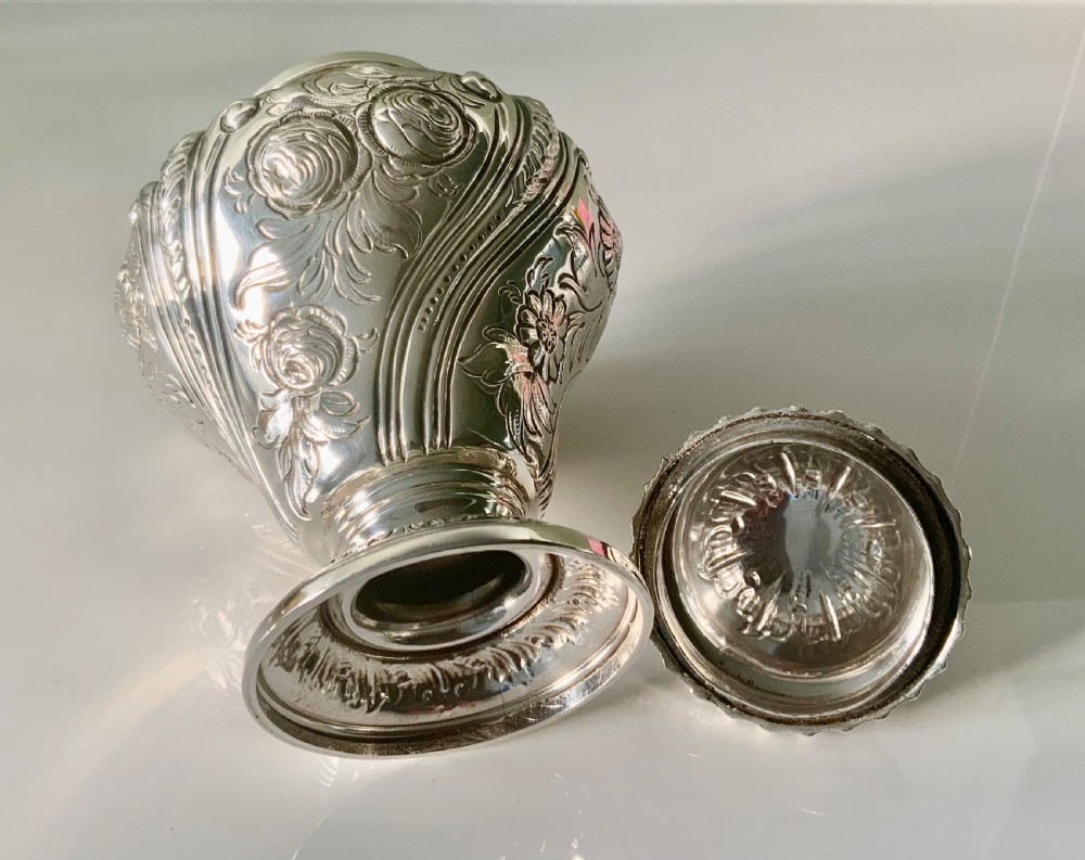 An ornate silver tea caddy featuring embossed scrollwork, a pedestal base, and a decorative finial on the lid. View X