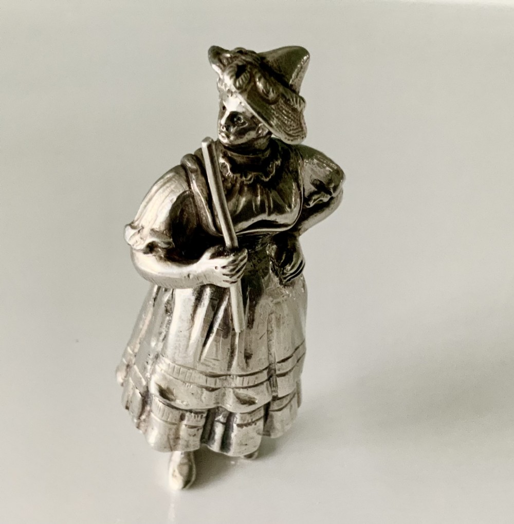 A cast silver perfume bottle sculpted in the form of a standing woman wearing a bonnet and dress while holding a staff.